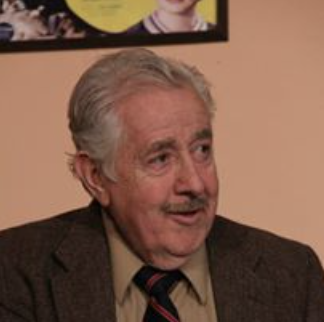 An elderly man with gray hair and a mustache, wearing a brown blazer, light shirt, and striped tie, sitting indoors with a beige wall in the background.