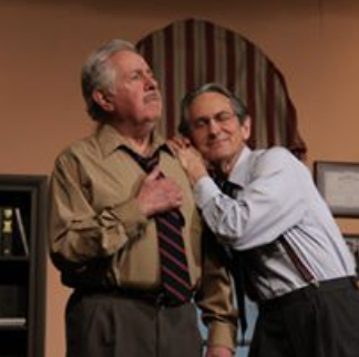 Two men, one older with gray hair and a beige shirt, the other younger with glasses and a white shirt, are in an office setting. The younger man is holding the older man's shoulder and appears to be comforting or consoling him.