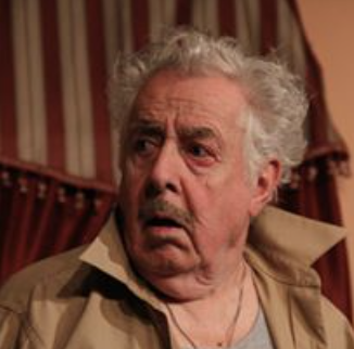 An elderly man with disheveled white hair and a surprised or shocked expression on his face, sitting indoors with striped wallpaper in the background.