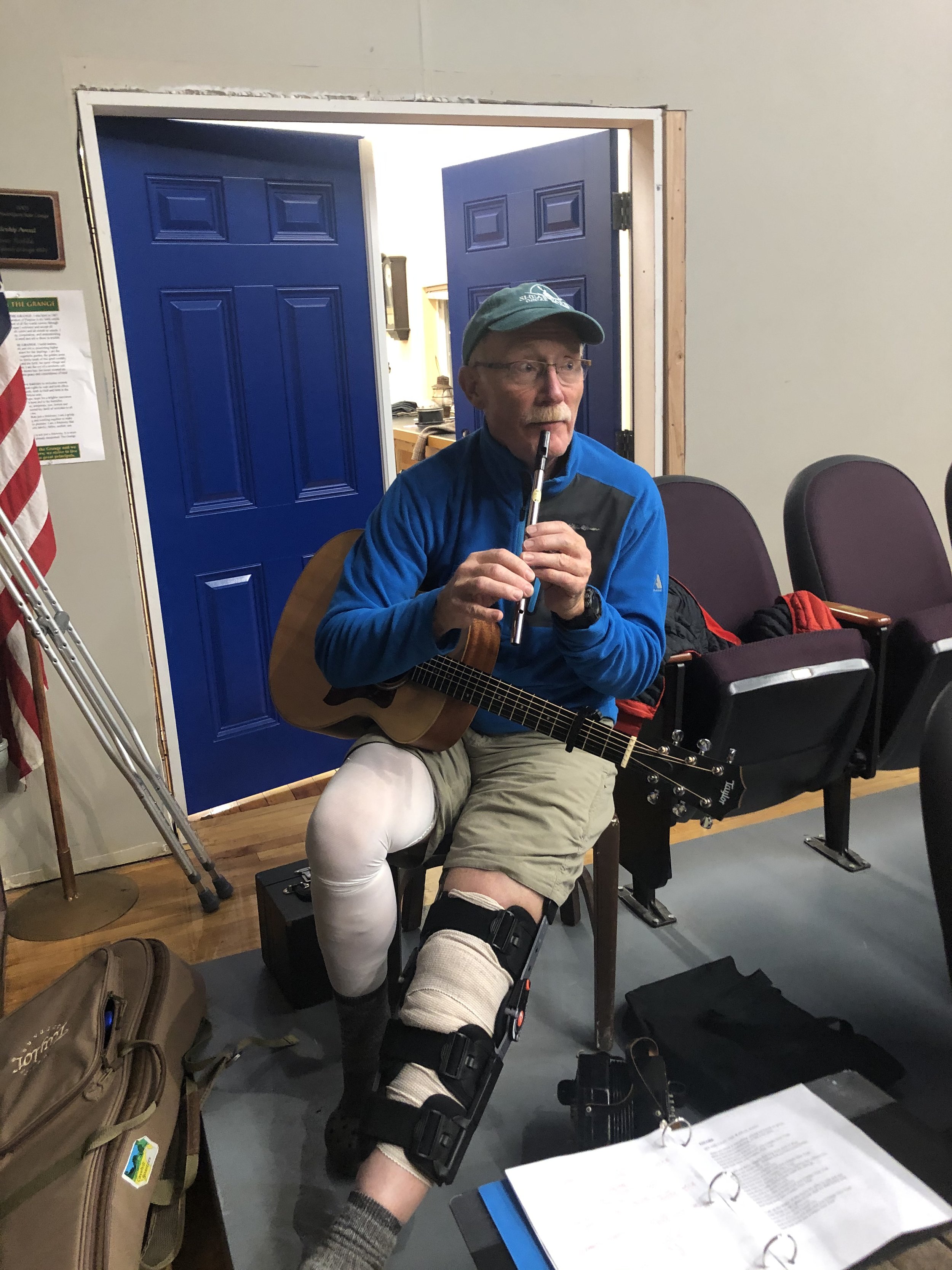 An elderly man playing a guitar and a harmonica, sitting on a chair in a room with purple seats, blue doors open to a room with a desk, wearing a knee brace, hat, and glasses.