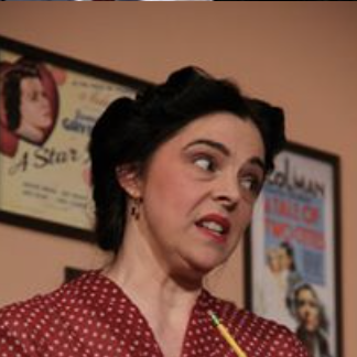 Woman with dark hair wearing a red polka dot dress, making a humorous facial expression in front of movie posters.