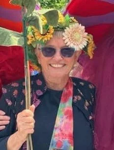 Smiling woman wearing sunglasses, a hat decorated with yellow and white flowers, and a colorful scarf, holding a rainbow umbrella.