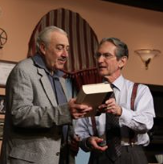 Two men, one elderly with white hair and the other younger, are standing indoors looking at a book together.