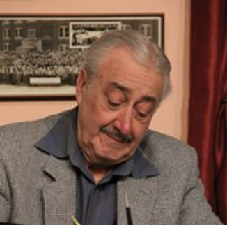 An elderly man with gray hair, wearing a gray blazer and a dark shirt, standing indoors with framed pictures on the wall behind him.