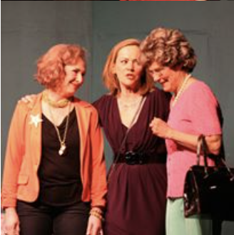Three women standing together and talking, one wearing an orange blazer, one in a dark dress, and one in a pink top with green pants, indoors.