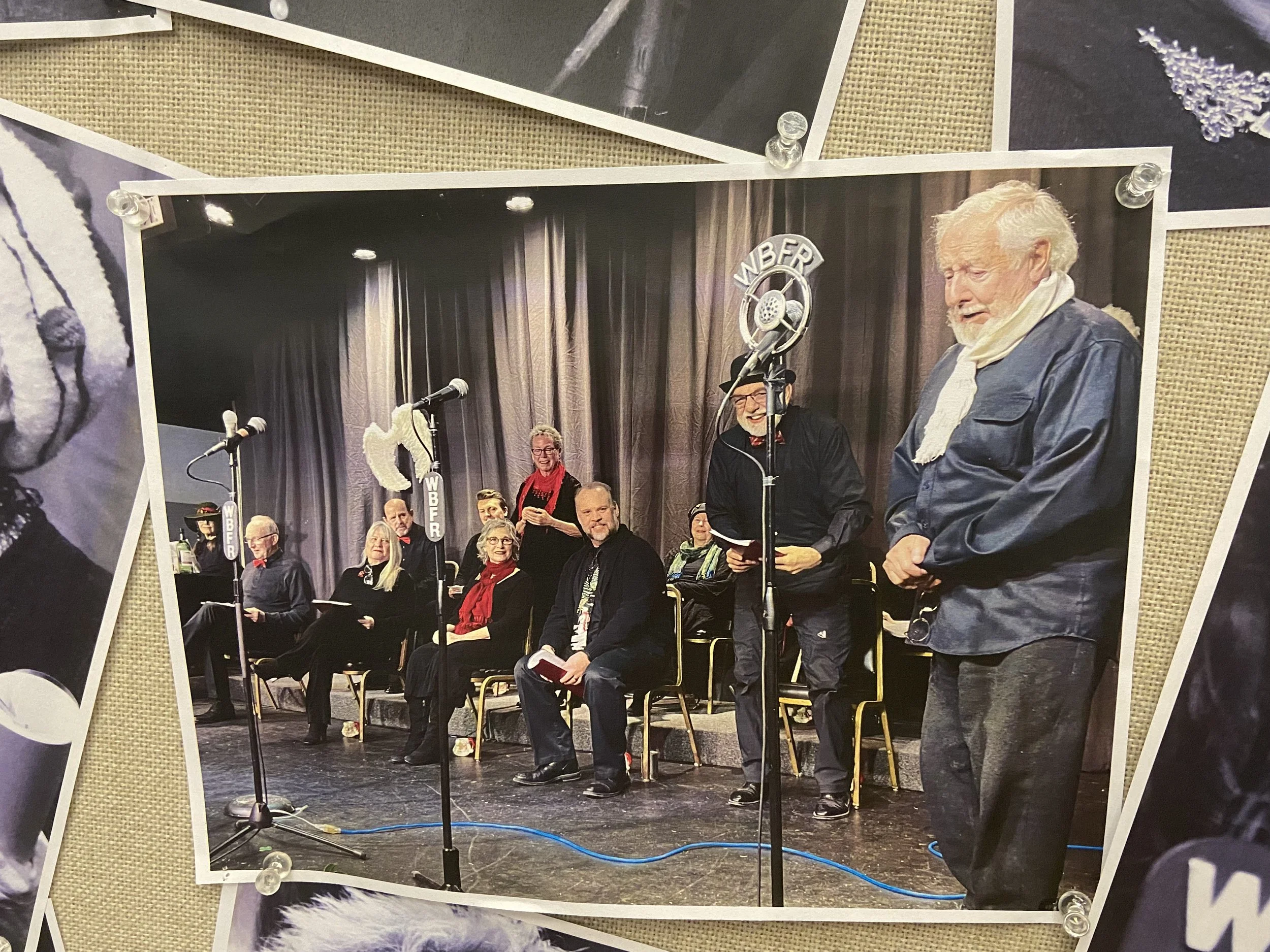 A group of people sitting and standing on stage during a radio broadcast with microphones and a 'WBER' sign, some holding scripts, others talking, in front of a curtain.