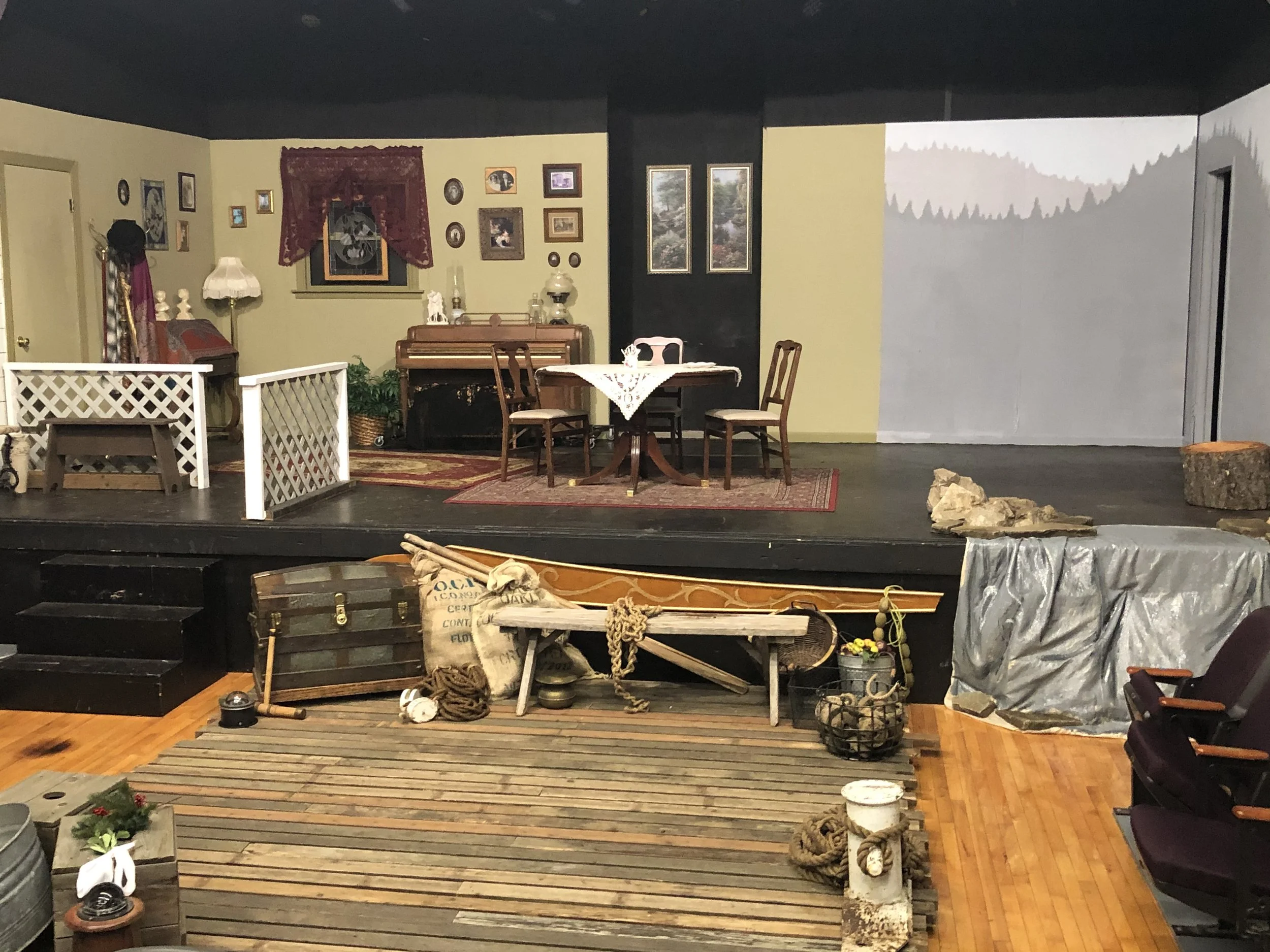 A stage set designed to resemble a cozy living room with a dining area, decorated with paintings, lamps, and furniture, and outdoor props like a canoe, logs, and farming tools in the foreground.