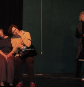 Two people sitting on a bench, one leaning on the other's shoulder, with a person standing nearby holding a microphone in a dark room.