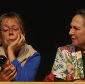 Two women sitting at a table, one appears upset and is looking at a phone, the other looks at her with concern.