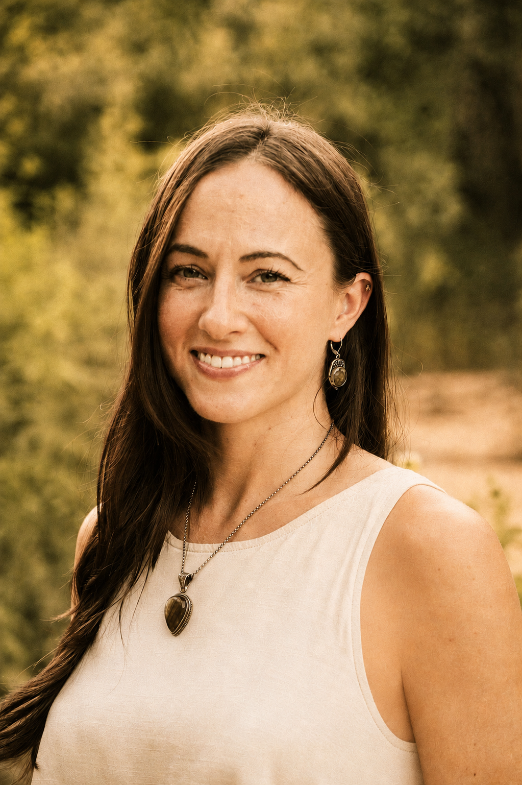 A woman with long brown hair smiling outdoors, wearing a white sleeveless top and jewelry, with green trees in the background.
