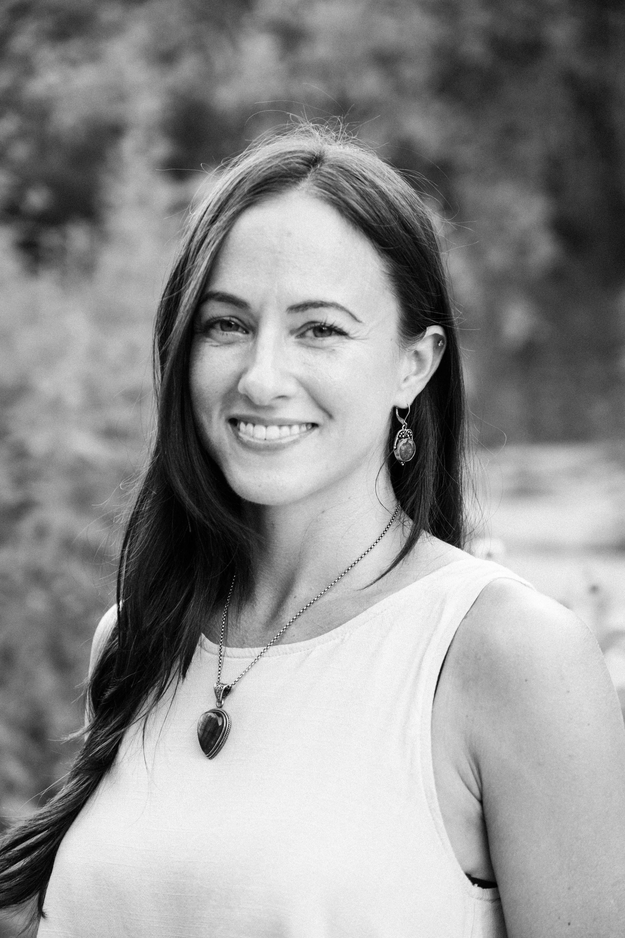 A woman with long dark hair smiling outdoors, wearing earrings, a necklace, and a sleeveless top.