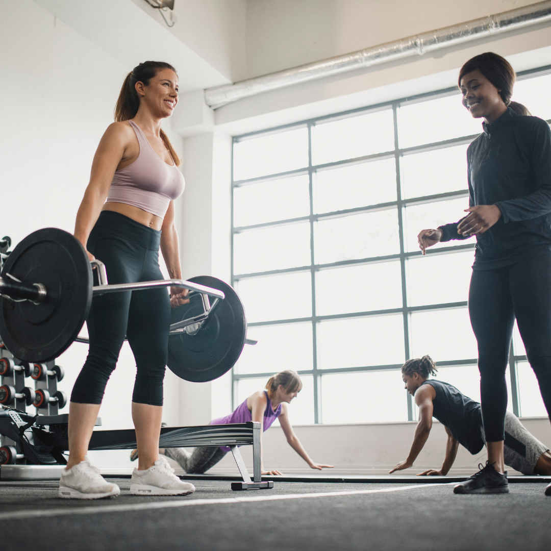 Women exercising in a gym with one lifting weights and others doing push-ups on the floor.
