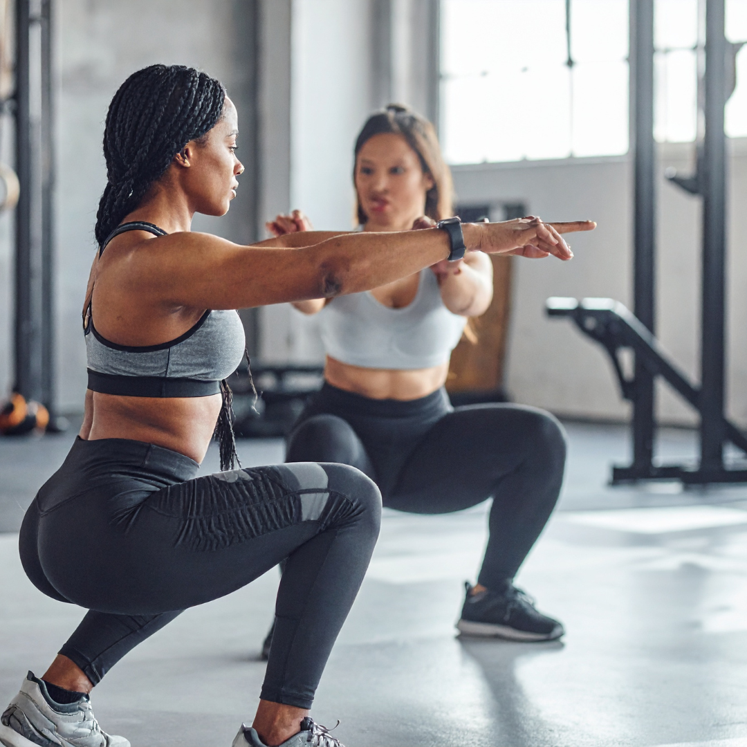 Two women in workout clothes squatting and stretching in gym, one in foreground with arms extended forward, and another behind them with arms stretching backward.