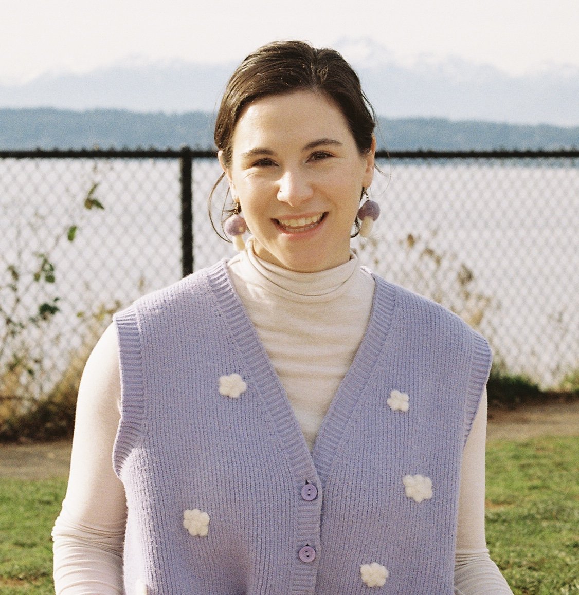 A woman smiling outdoors near a chain-link fence with mountains in the background, wearing a light purple knitted vest with white flower patterns over a cream turtleneck.
