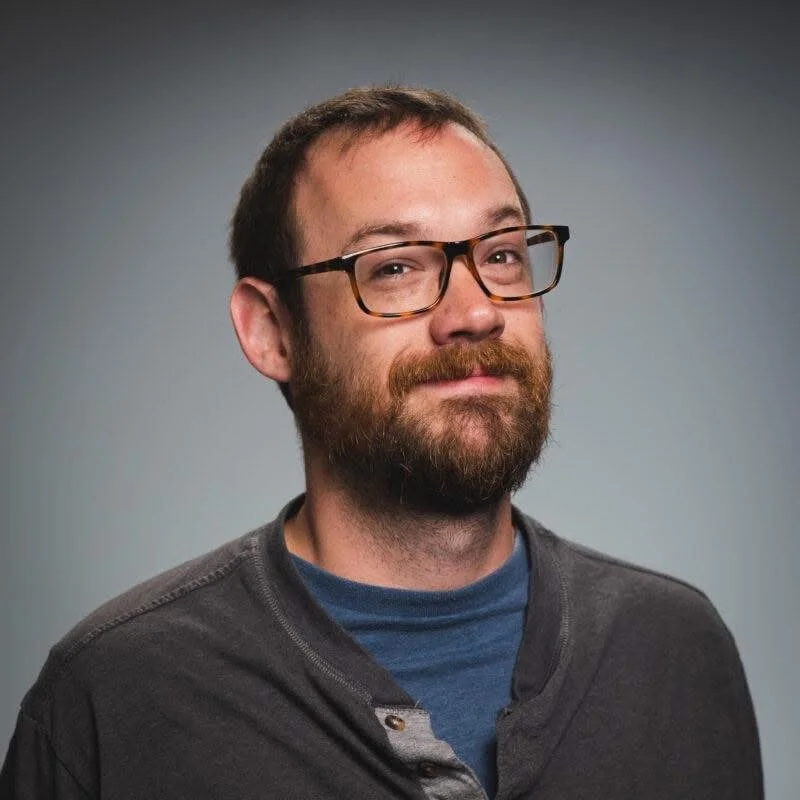 A man with glasses, a beard, and short brown hair, dressed in a dark shirt and blue shirt underneath, looking at the camera with a slight smile against a gray background.
