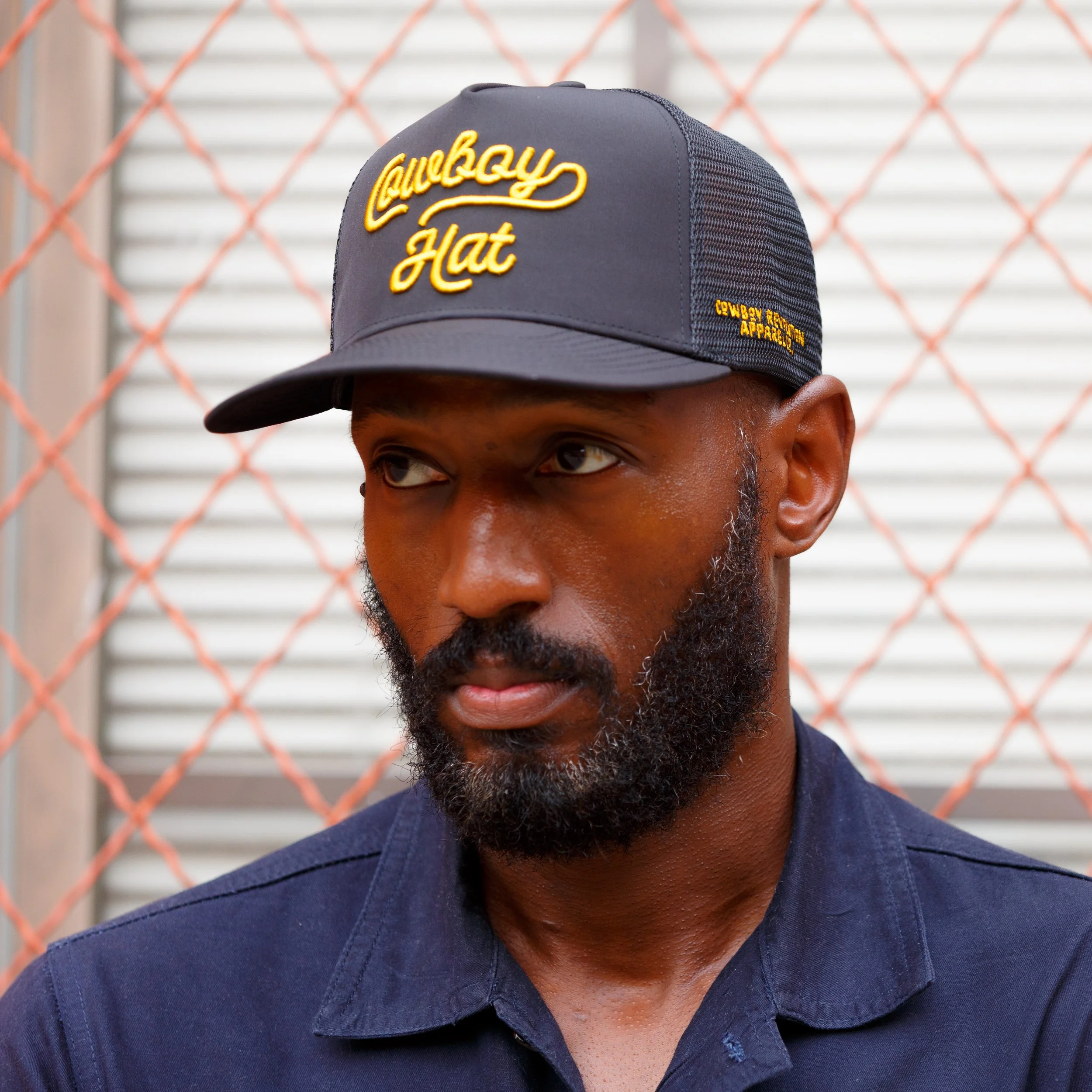 A man with a beard wearing a navy blue shirt and a navy baseball cap with yellow embroidery reading 'Cowboy Hat' and 'Cowboy Rides and Accessories'. He is standing outdoors in front of a chain-link fence and a building with metal siding.