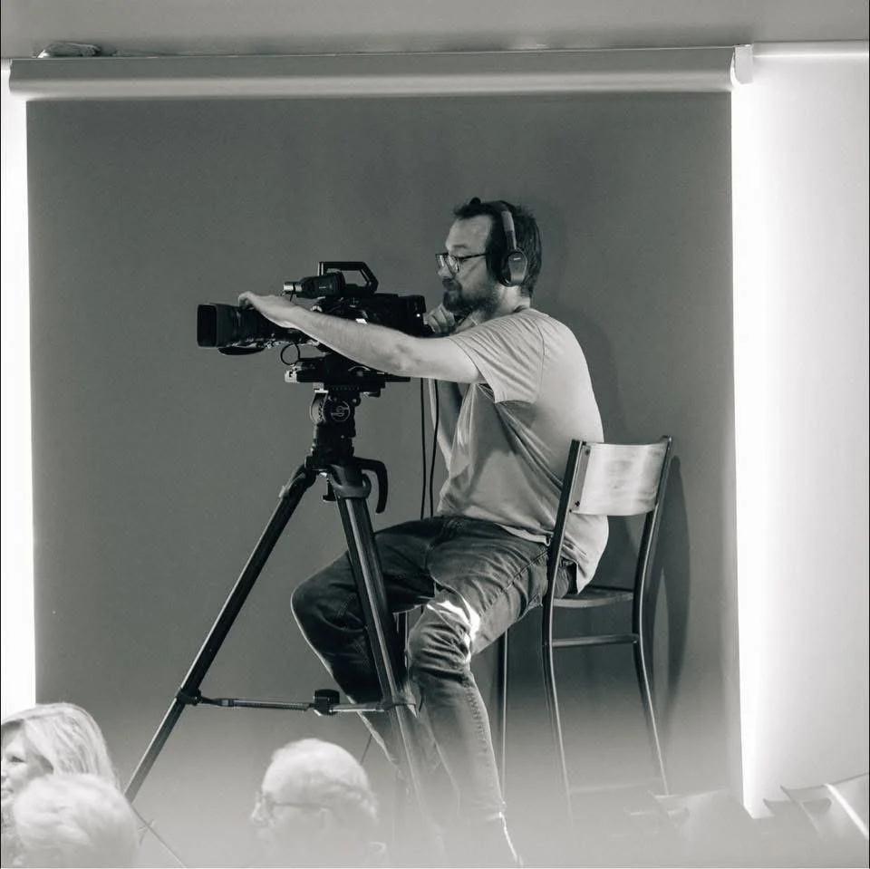 A man operating a professional video camera on a tripod, sitting on a chair, wearing headphones and glasses, in front of a plain wall.