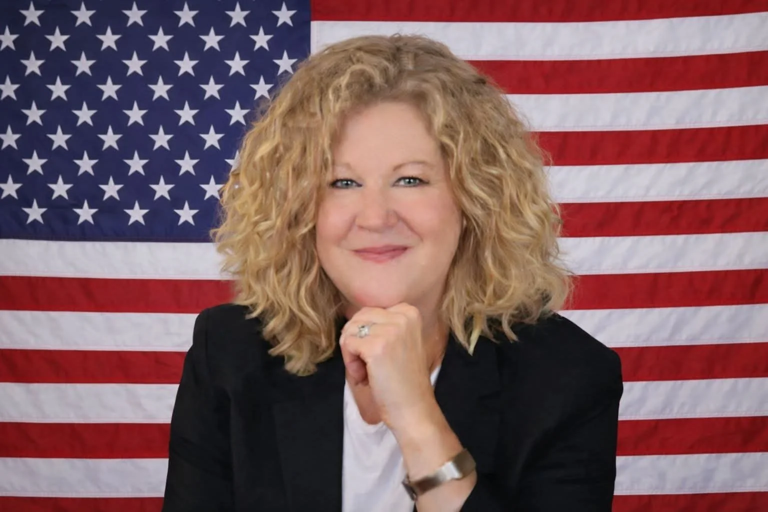 A woman with curly blonde hair, wearing a black blazer and white top, posing in front of an American flag with stars and stripes.
