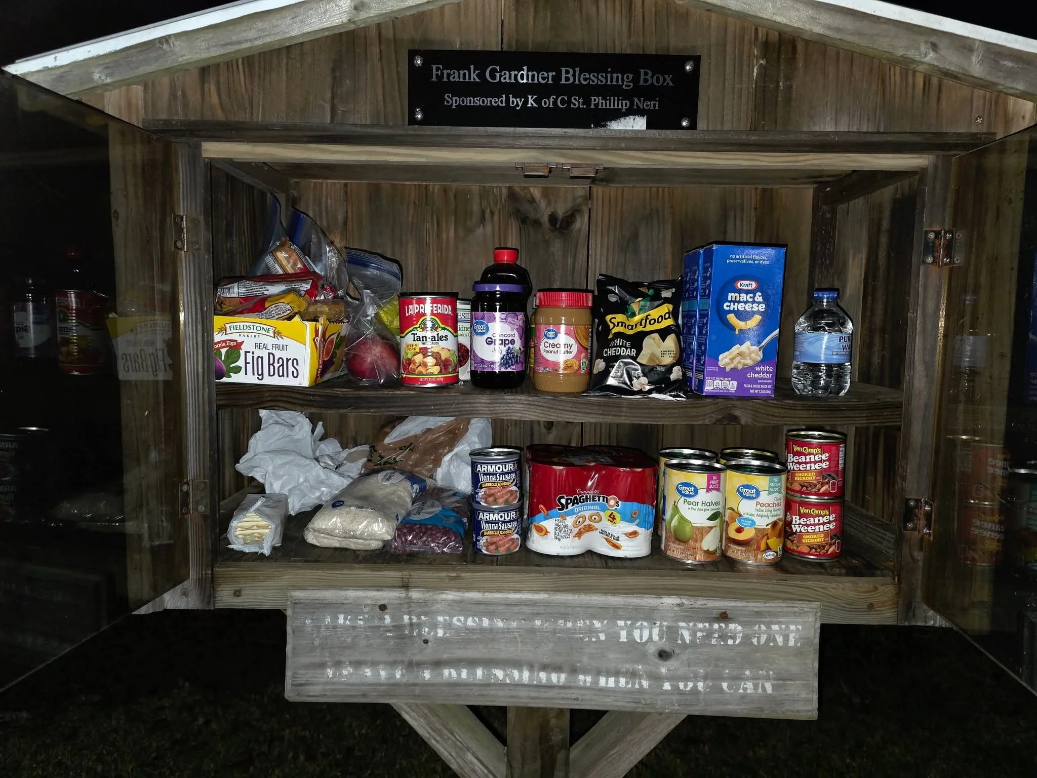 Inside a wooden blessing box cabinet with various food items including fig bars, canned meats, pasta, juice, and bottled water, with a sign on top reading 'Frank Gardner Blessing Box'.