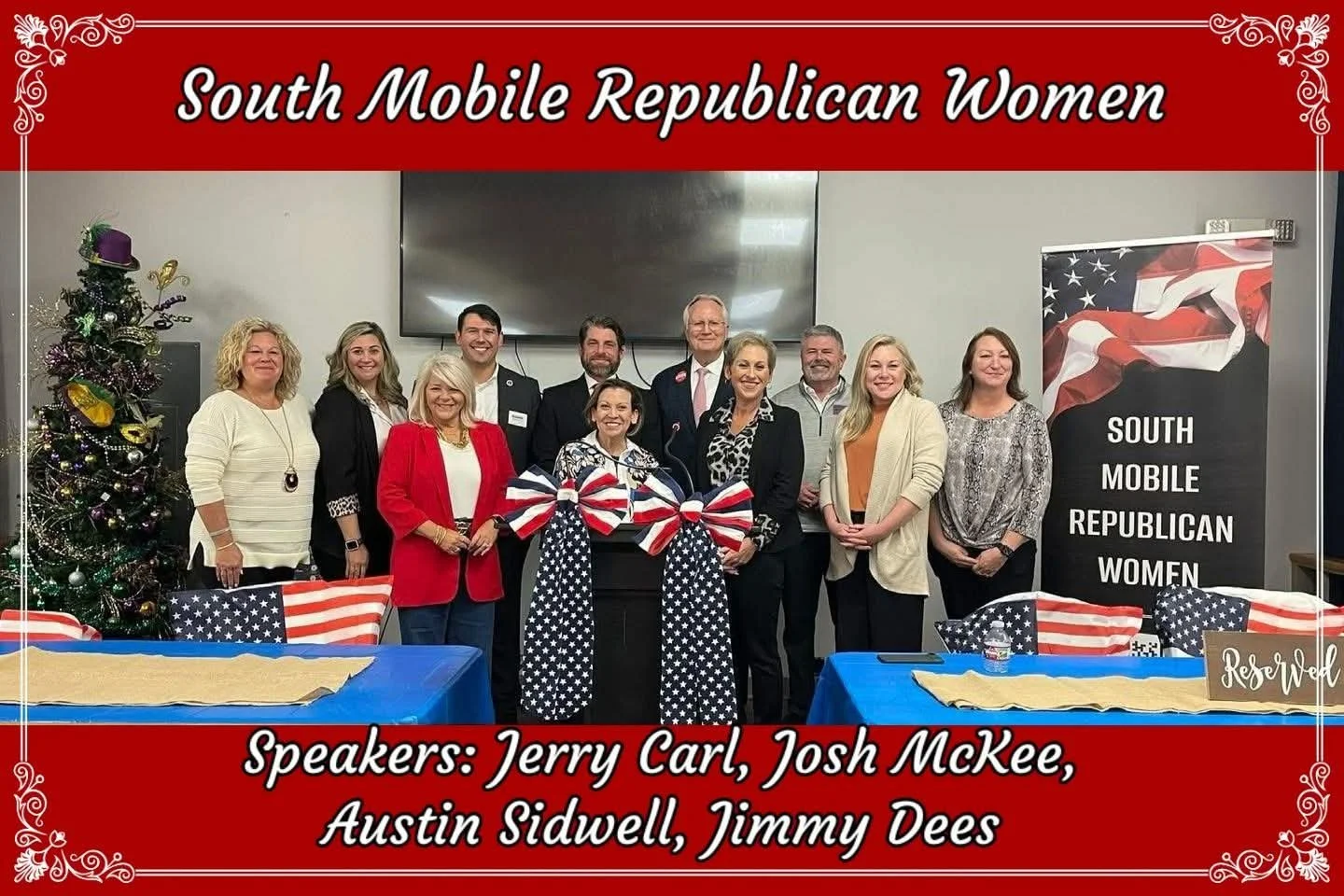 A group of women and men gathered for a South Mobile Republican Women event, standing behind decorated tables with American flags and a Christmas tree in the background.