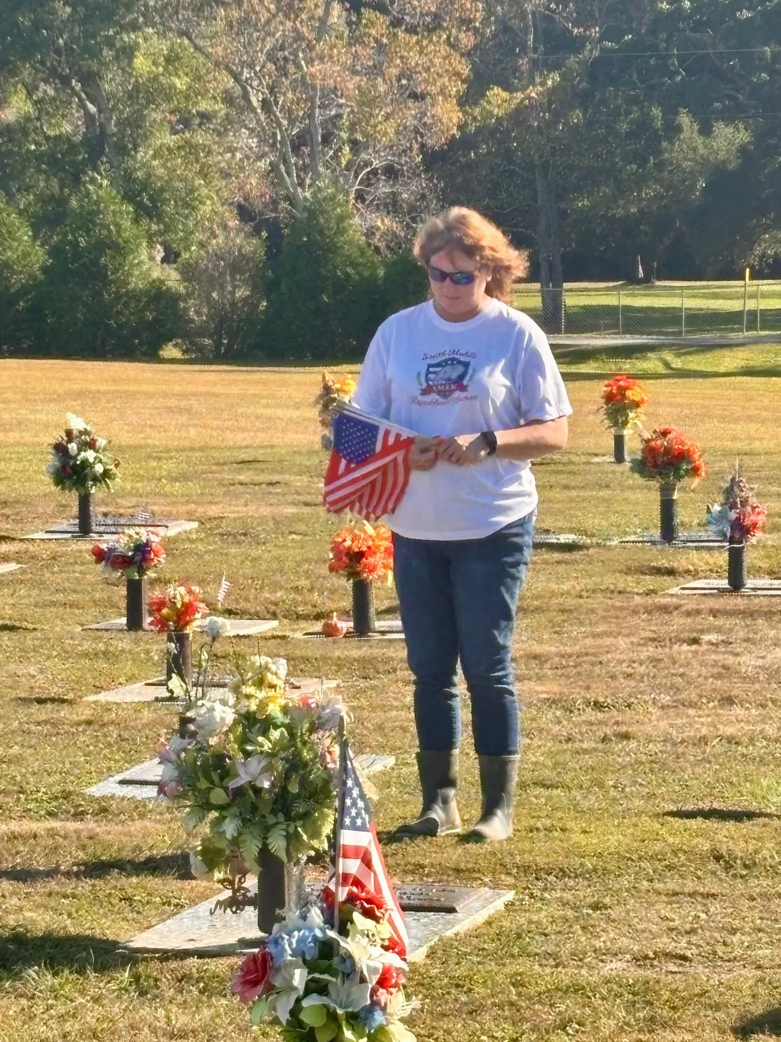 Woman in a white T-shirt, jeans, and boots walking through a cemetery decorated with flowers and flags, holding American flags in her hands.