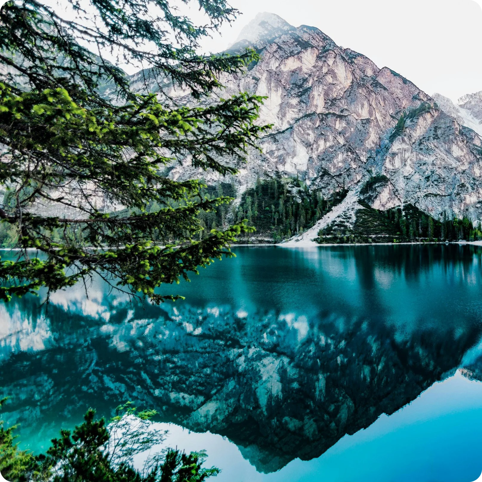 A mountain with rocky cliffs and evergreen trees next to a calm, turquoise lake, reflected in the water.