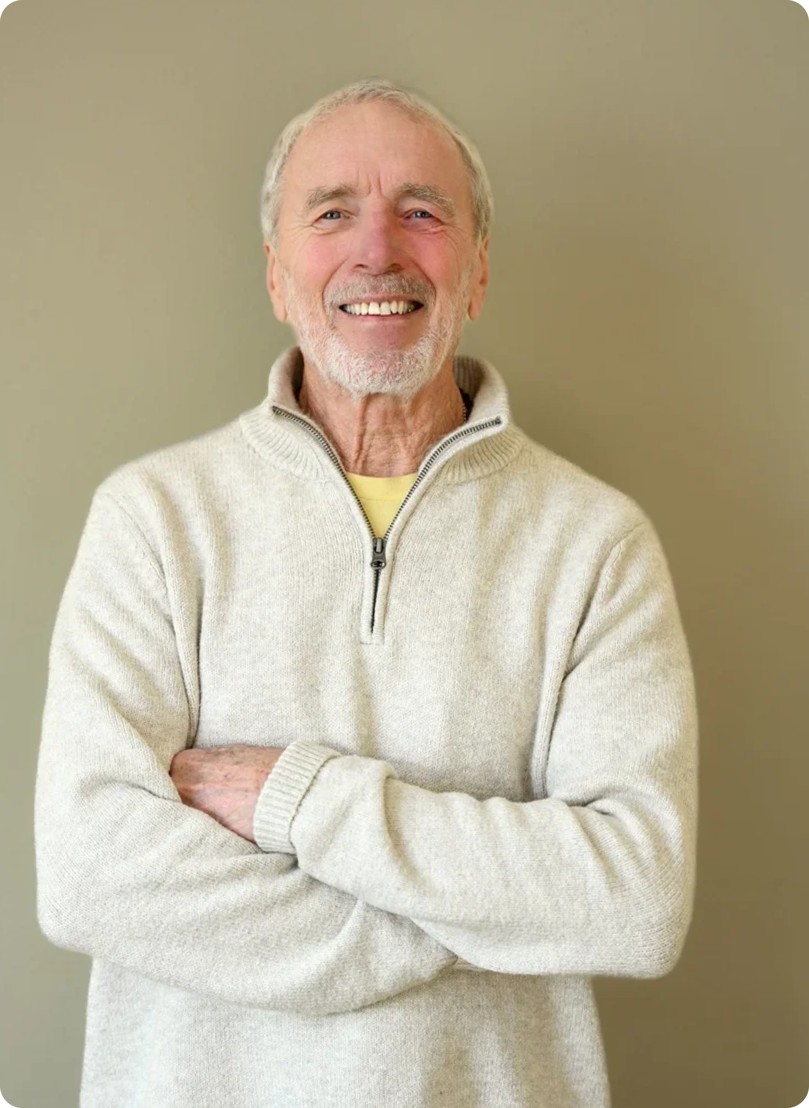 Close-up of a smiling older man with gray hair and beard, wearing a cream-colored zip-up sweater with arms crossed, standing against a plain beige background.