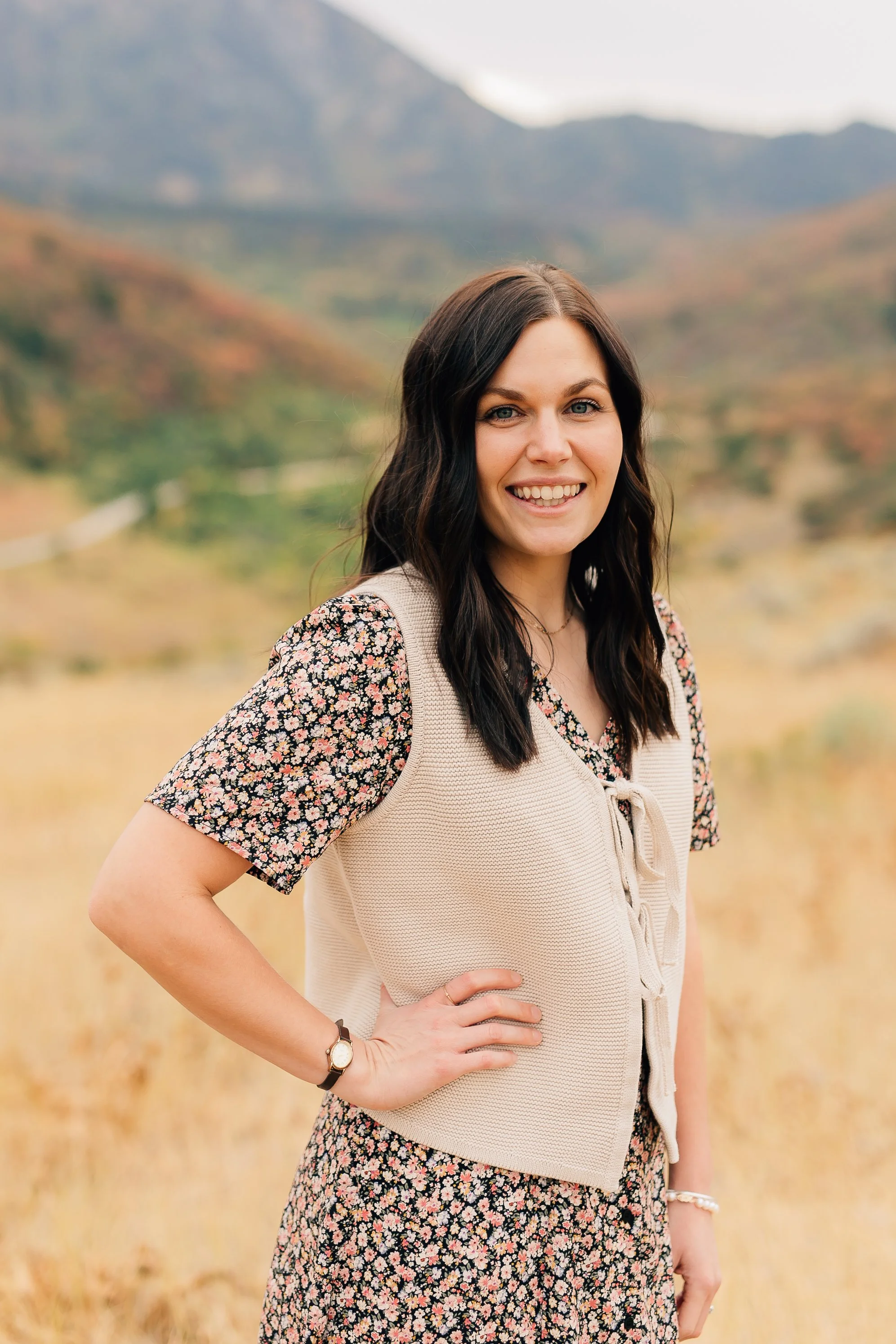 A woman with long dark hair, wearing a floral dress and a light-colored vest, standing outdoors in a field with mountains in the background.
