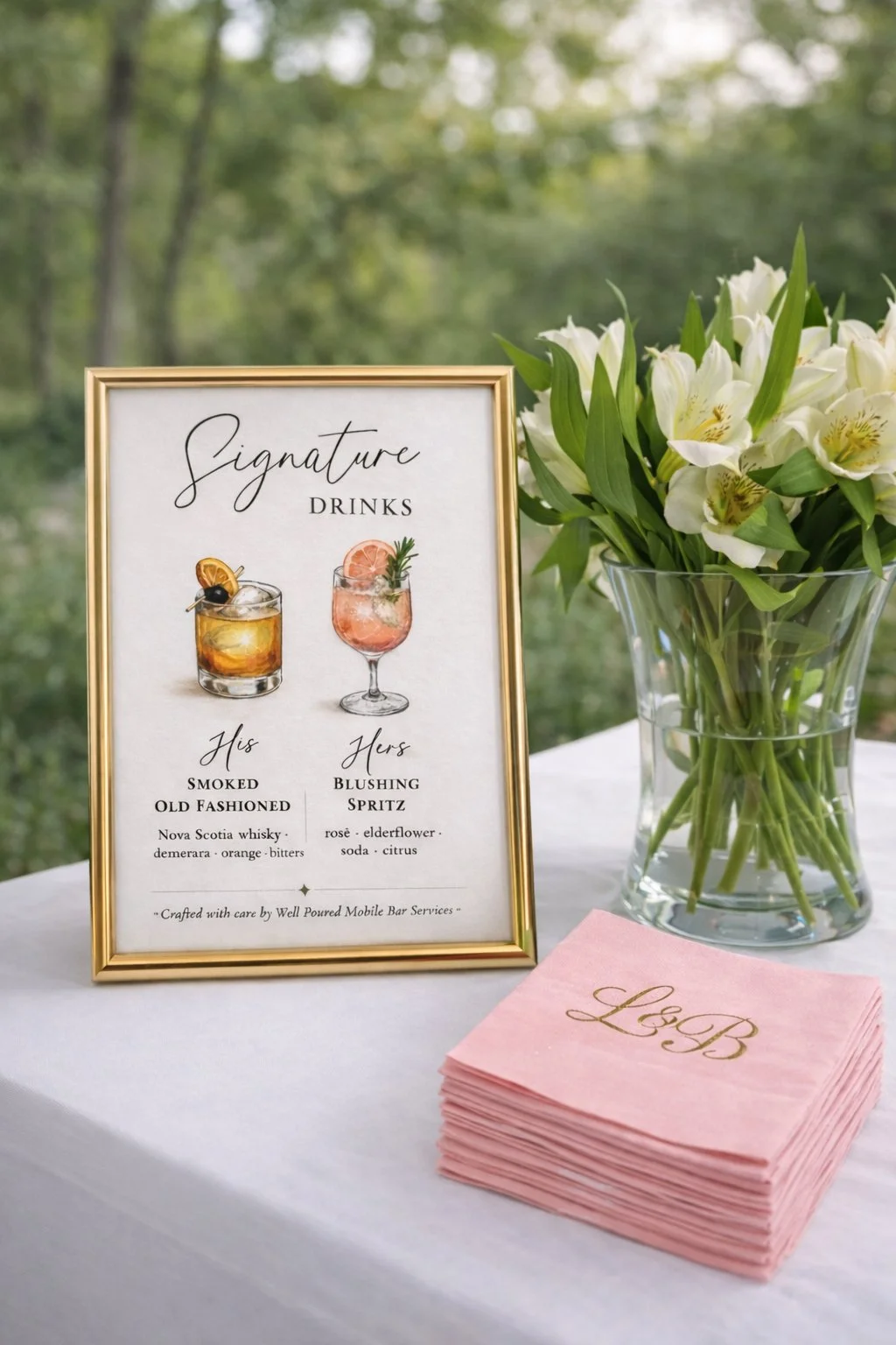 A framed drink menu featuring signature cocktails with illustrations, a vase of white flowers, and a stack of pink napkins with gold monogram initials on a table outdoors.