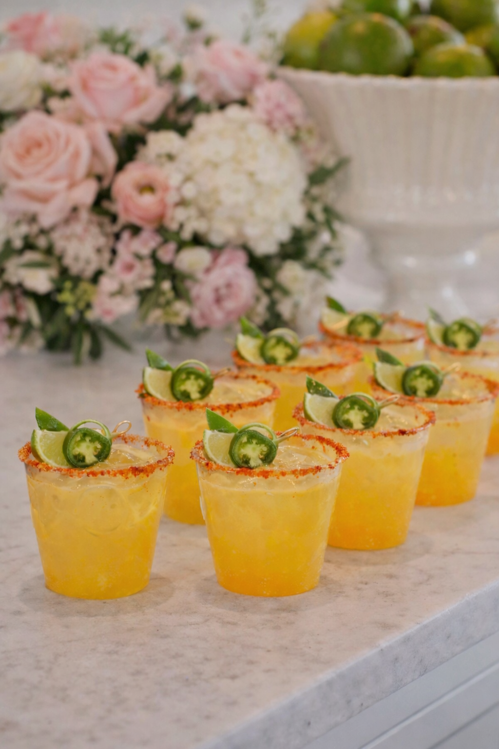 Six yellow cocktails garnished with lime wedges and jalapeño slices on a white surface, with a backdrop of pink and white flowers and a bowl of green fruit.