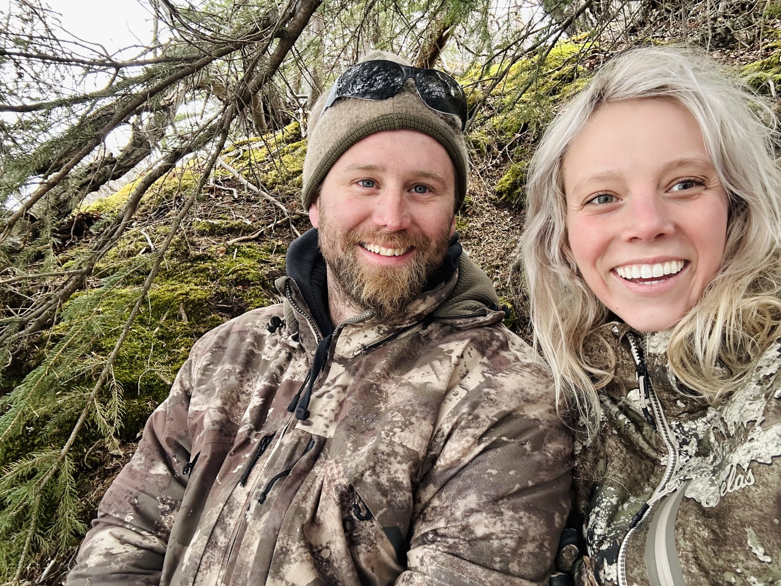 A smiling man and woman in camouflage jackets outdoors in a wooded area, with branches and moss in the background.