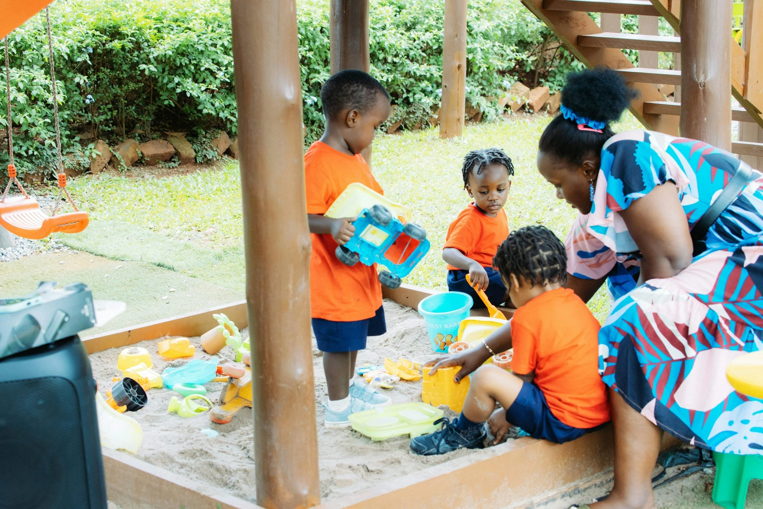 A woman and four children playing in a sandbox outdoors, surrounded by green bushes and trees. The children are wearing orange shirts and playing with sand toys, while the woman is engaging with them.