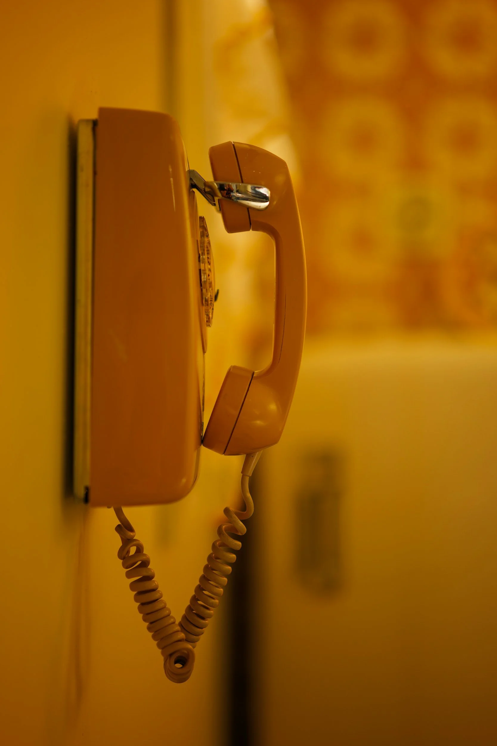 A vintage beige rotary phone mounted on a yellow wall, with its cord hanging down.