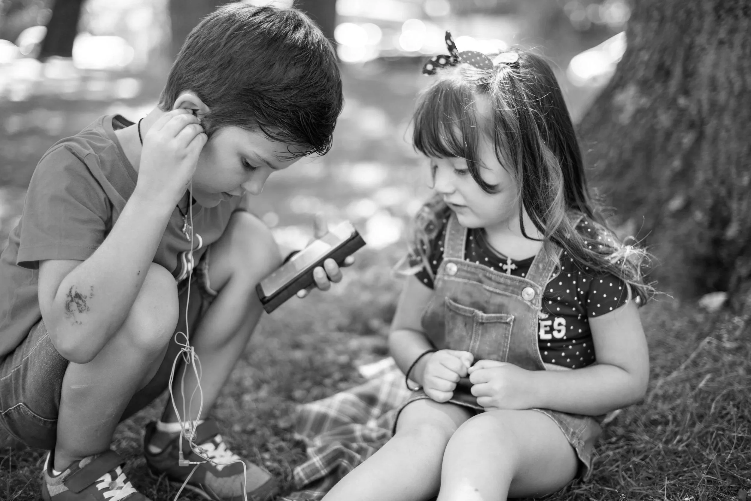 A young boy squatting on the ground looking at a smartphone, with headphones in his ears, alongside a young girl sitting on the grass near a tree, wearing overalls and a polka dot shirt, both outdoors in a park or forest setting.
