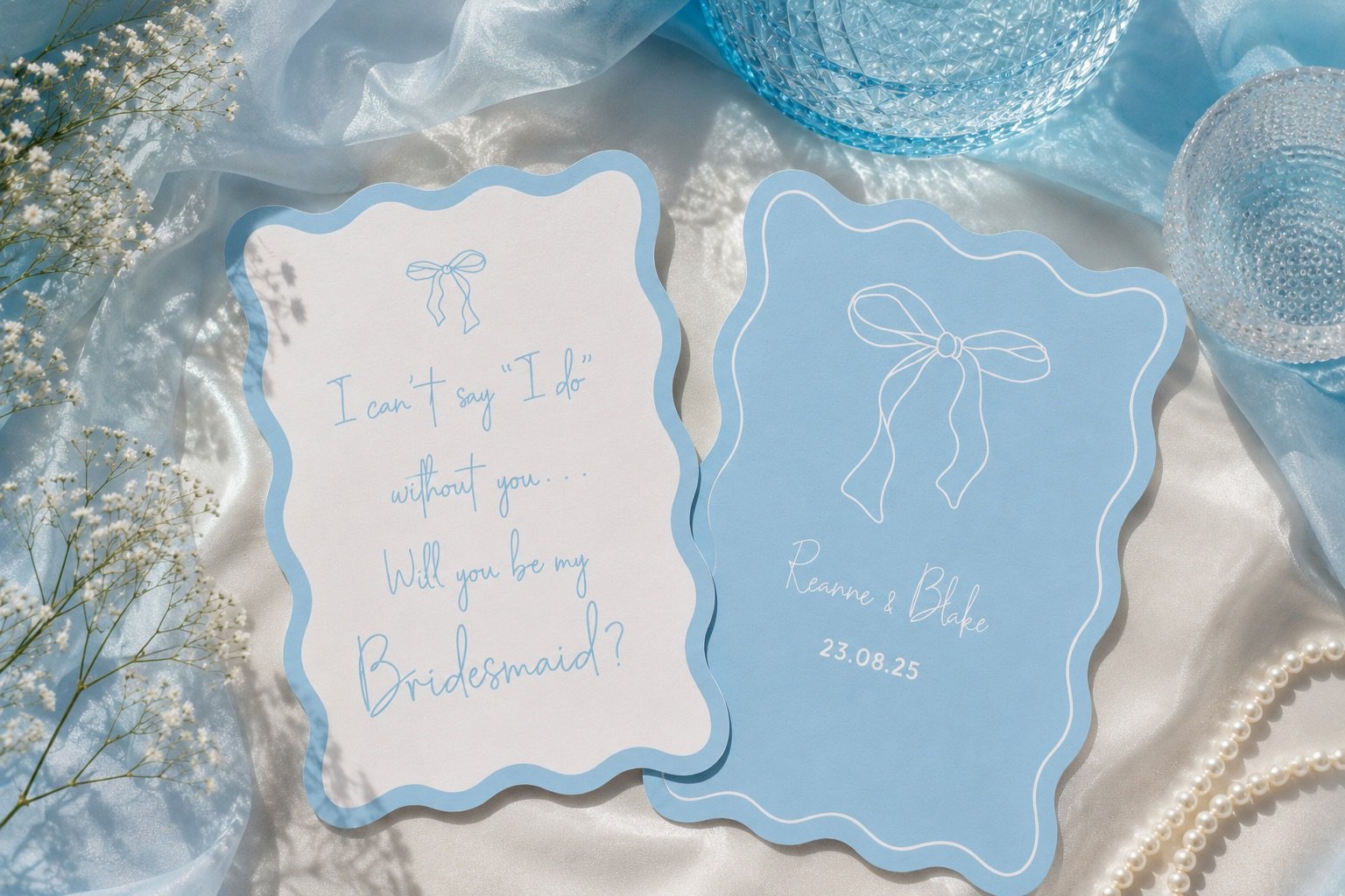 Two light blue wedding cards with white borders, one with a quote and the other with names and date, surrounded by white flowers, pearls, and blue vases on a silky fabric background.