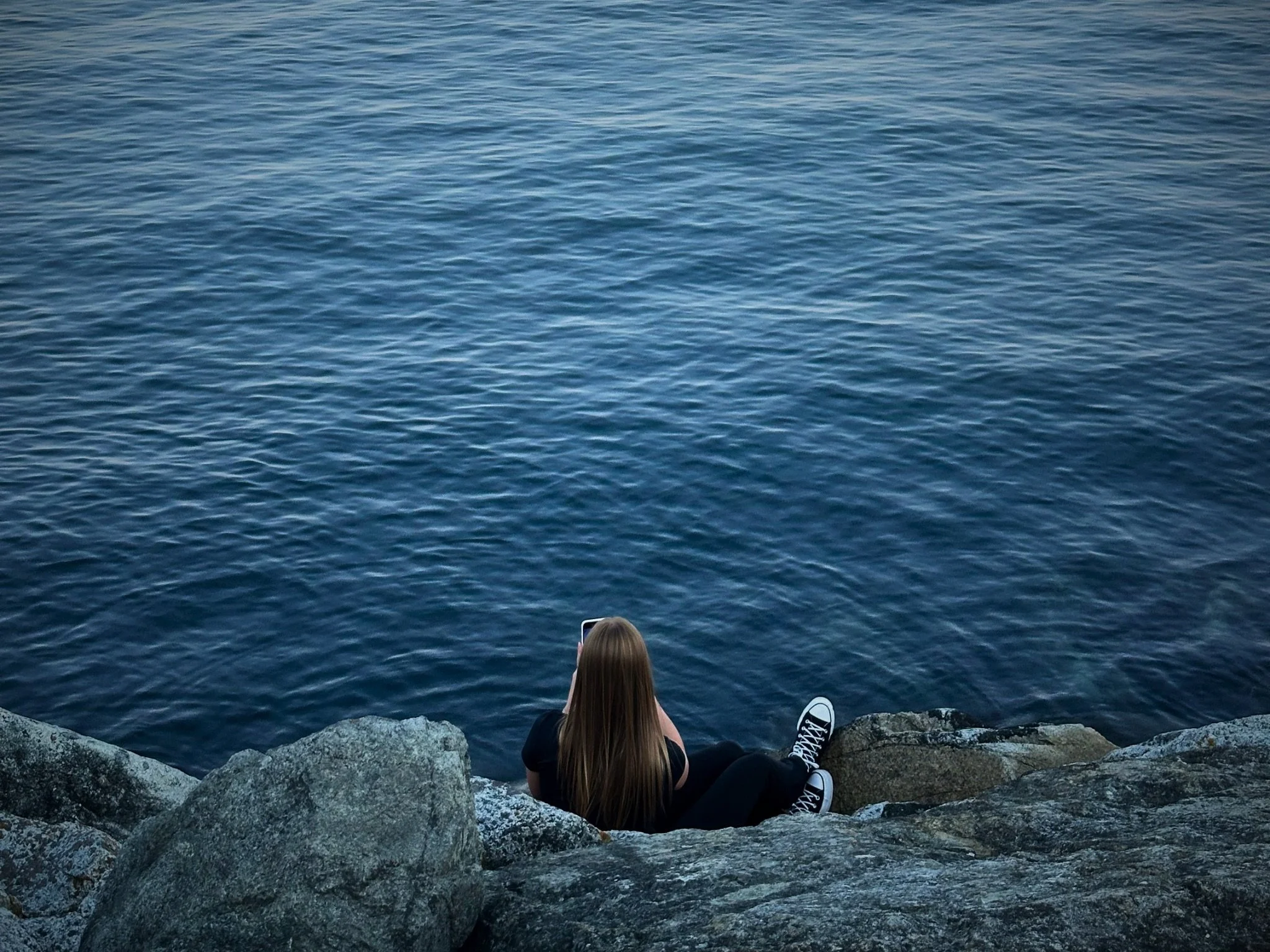 A woman with long hair sitting on rocks by the water, looking out at the blue ocean with small waves.