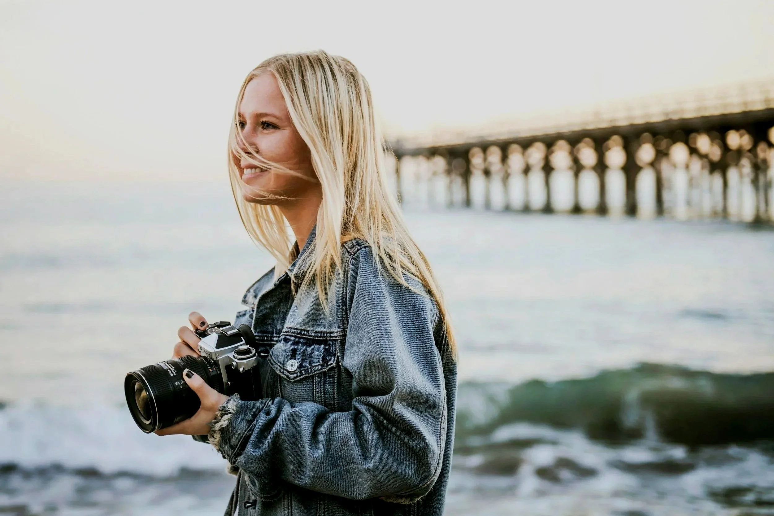 A woman with blonde hair holding a camera on the beach near the ocean with a pier in the background.