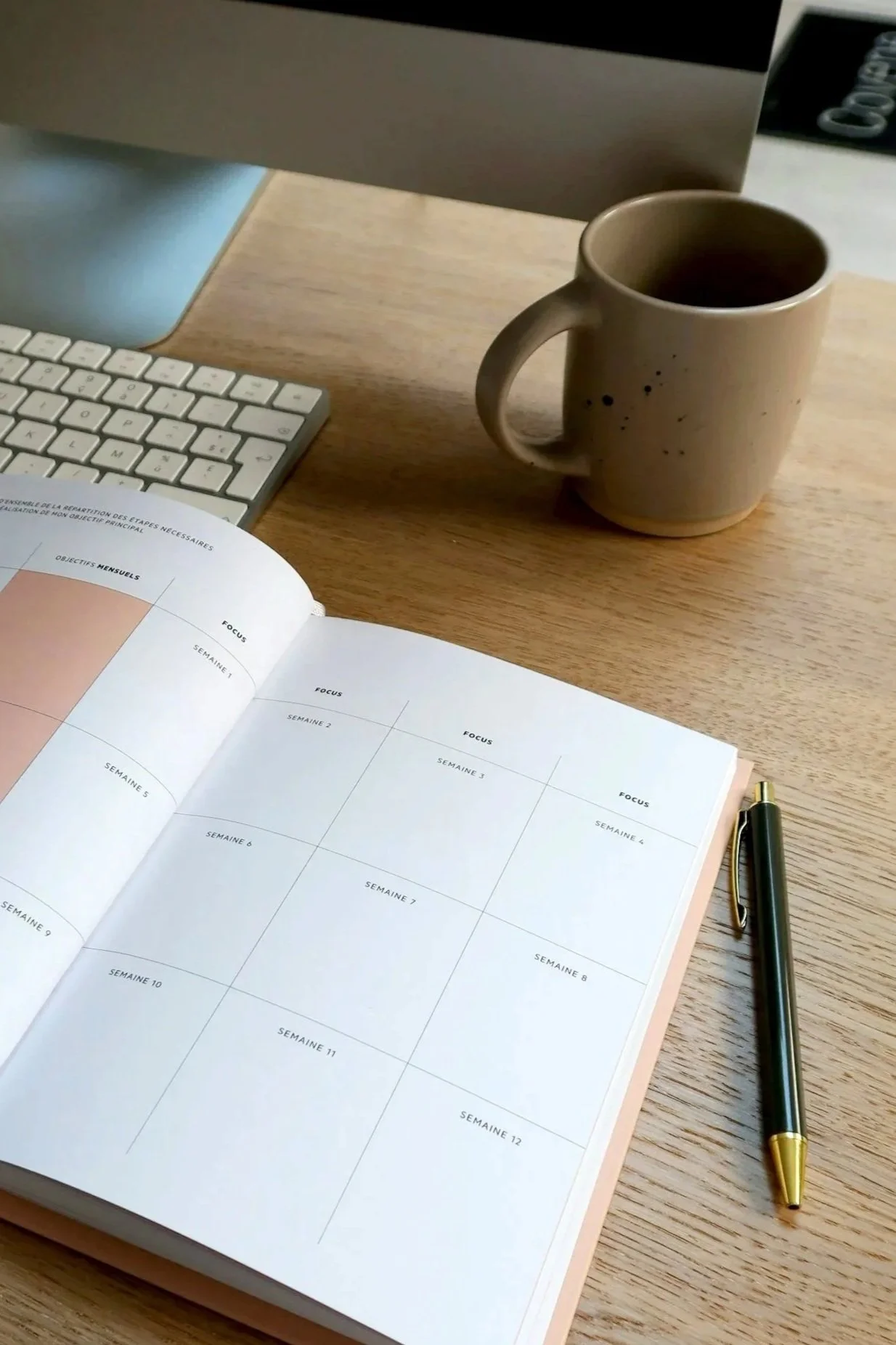 A desk with a computer monitor, a white keyboard, a tan mug with a hot drink, an open planner with weekly sections, and a black and gold pen.