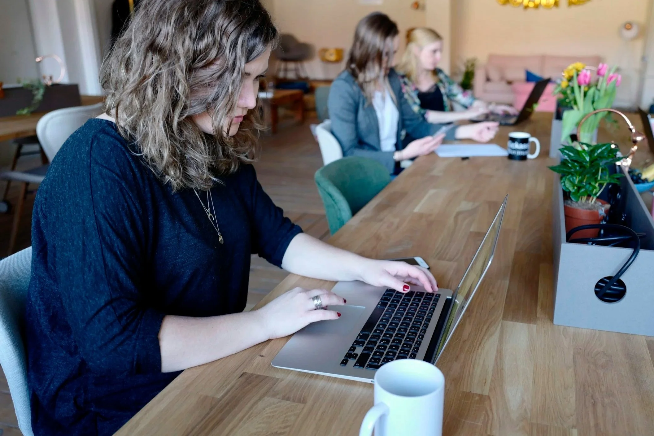 Women working on laptops and tablets at a long wooden table in a bright, modern office with plants and flowers.
