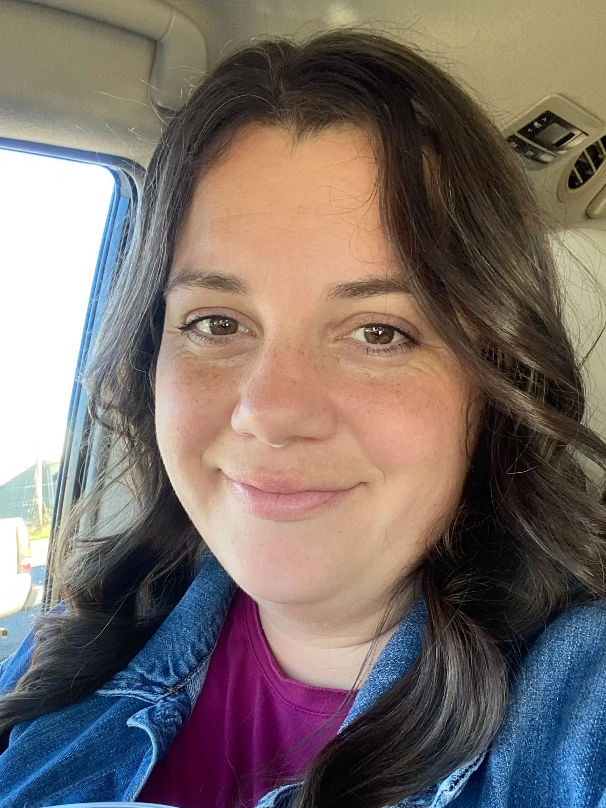 Close-up of a woman with brown hair and clear skin smiling, inside a vehicle.