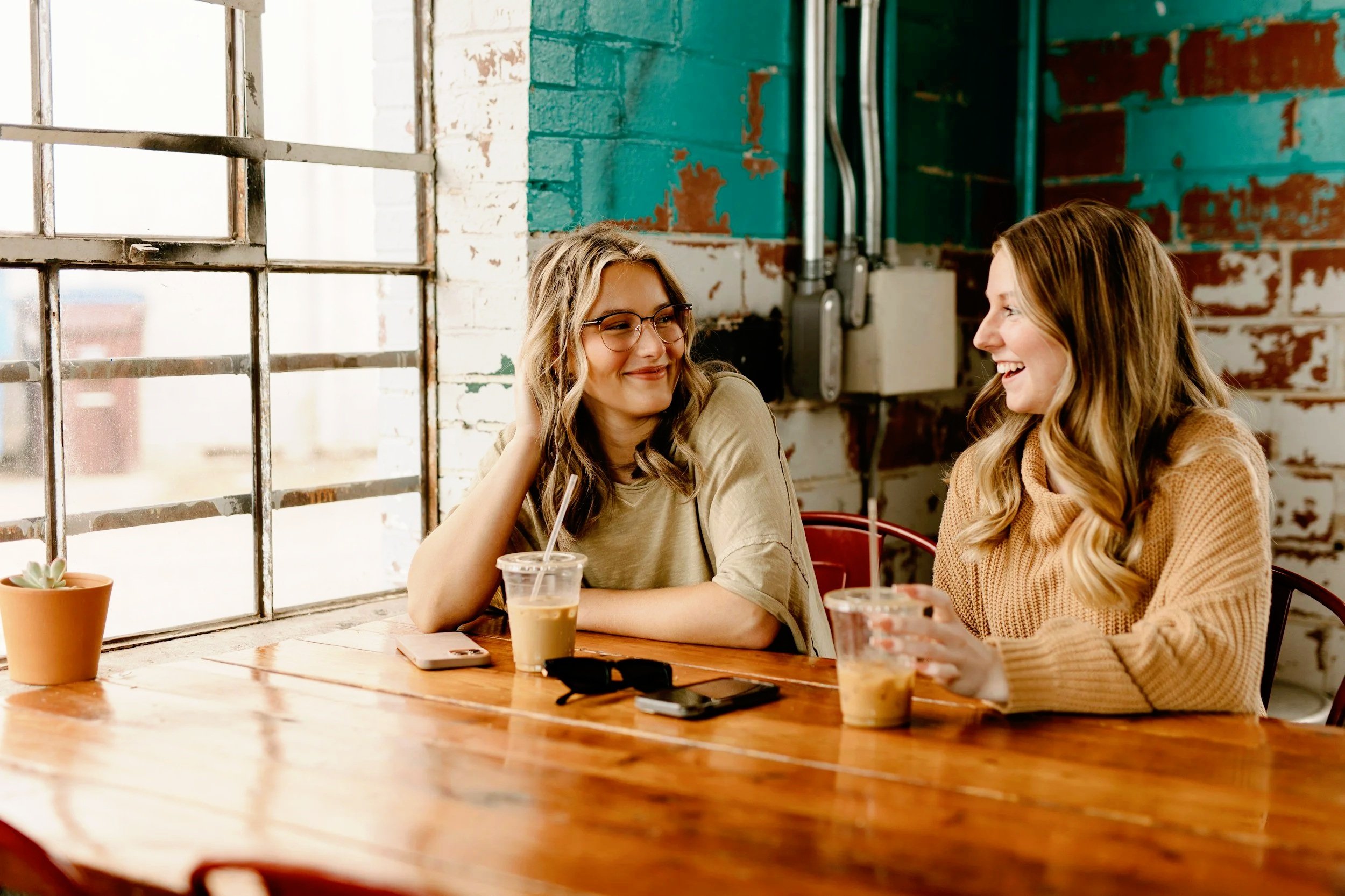Two young women sitting at a wooden table, smiling and talking in a coffee shop with a rustic interior, large window, and potted succulent plant.