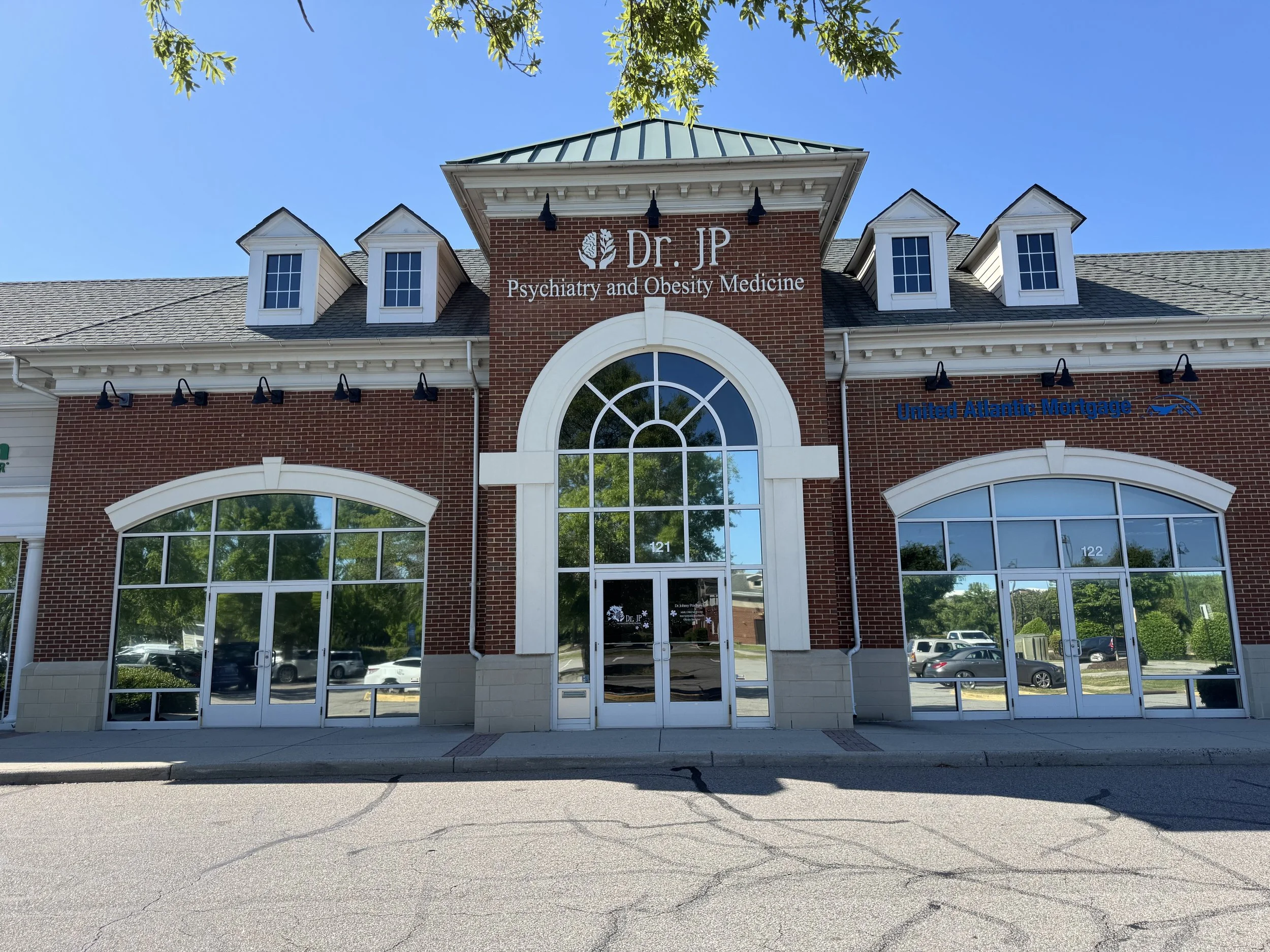 A brick and white building with large glass windows, housing a psychiatry and obesity medicine clinic, with a blue sky and green trees overhead.
