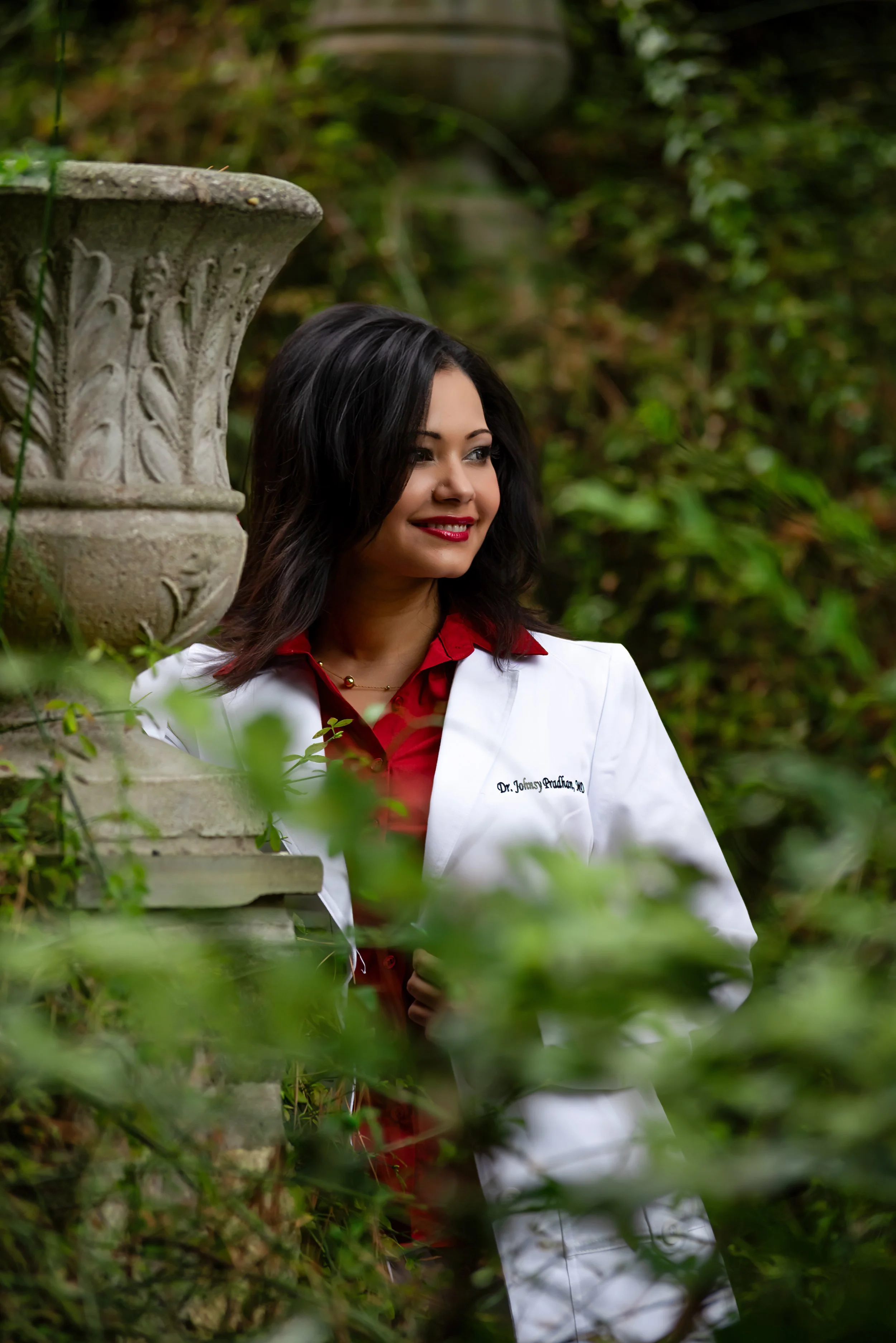 A woman with dark hair, wearing a white coat and red shirt, stands in a garden with greenery and an ornate stone planter nearby. She has a slight smile and looks to the side.