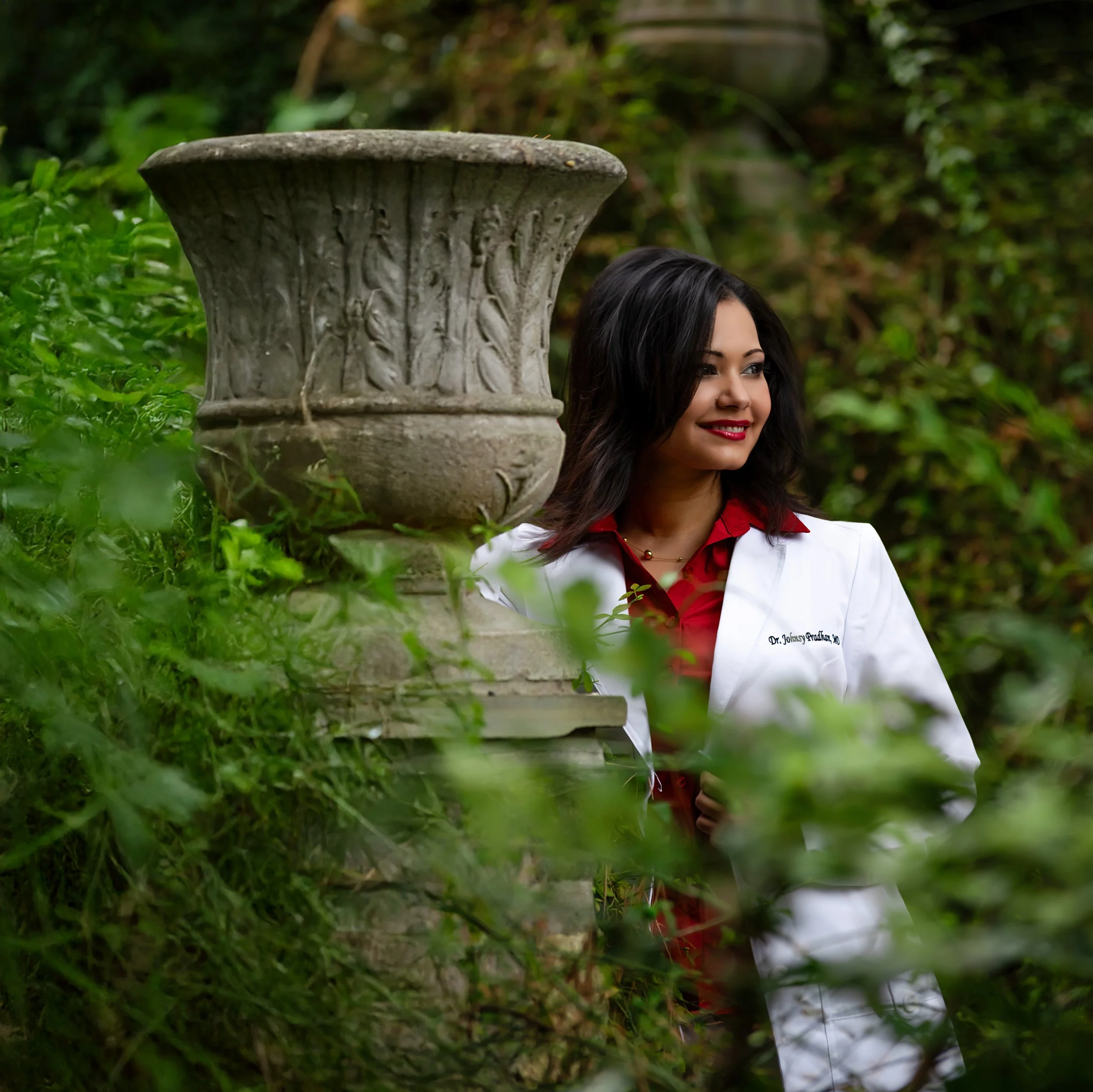 A woman with dark hair wearing a white coat and red shirt stands in greenery near a decorative stone urn.
