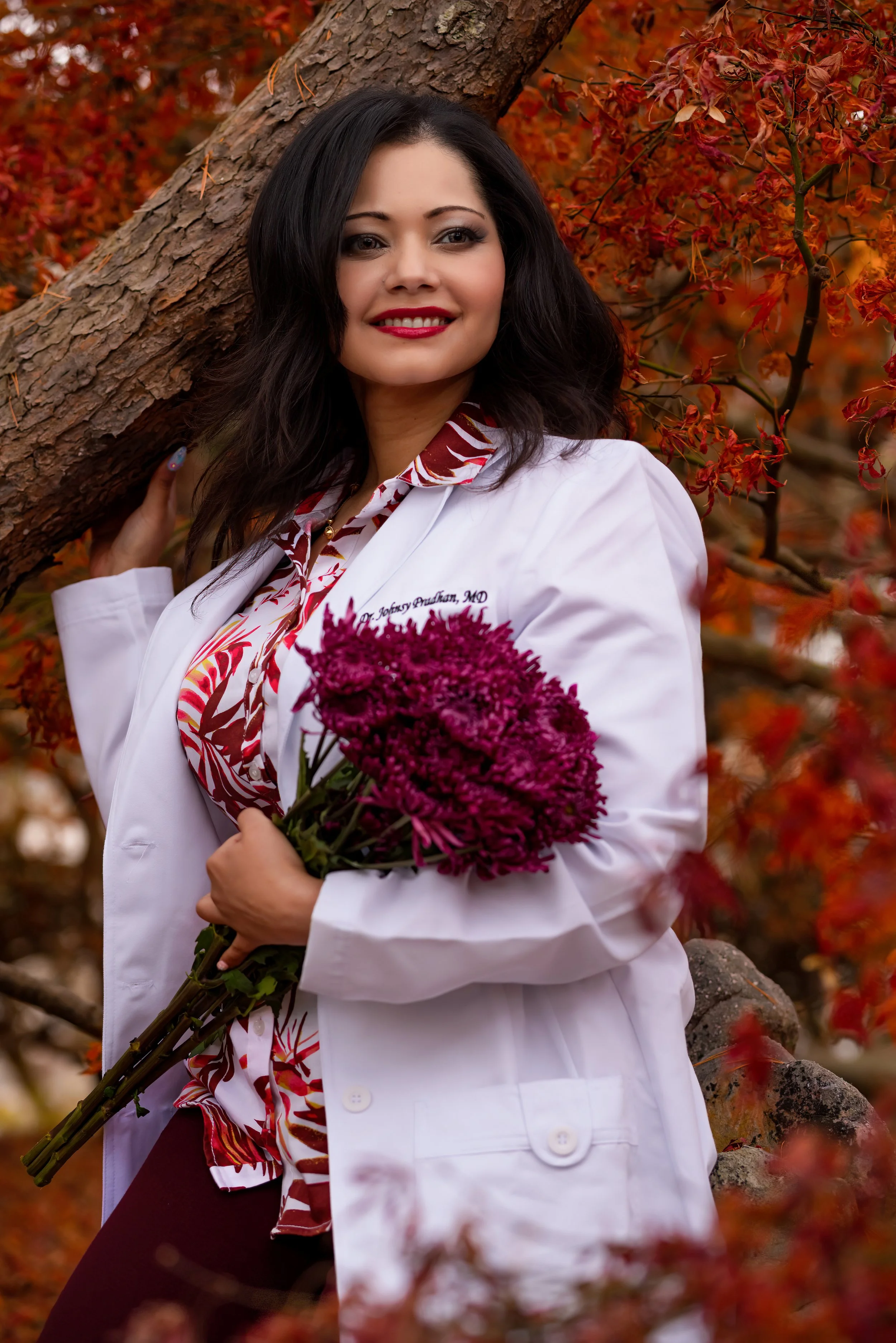 A woman in a white coat holding a bouquet of purple flowers outdoors during autumn, with red and orange leaves in the background.