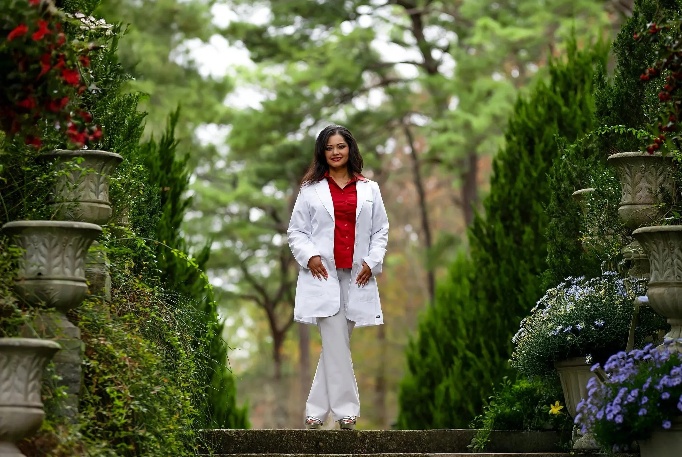A woman in a white doctor's coat and red shirt standing outdoors on a stone pathway, surrounded by green bushes and trees, with potted plants with flowers on either side.