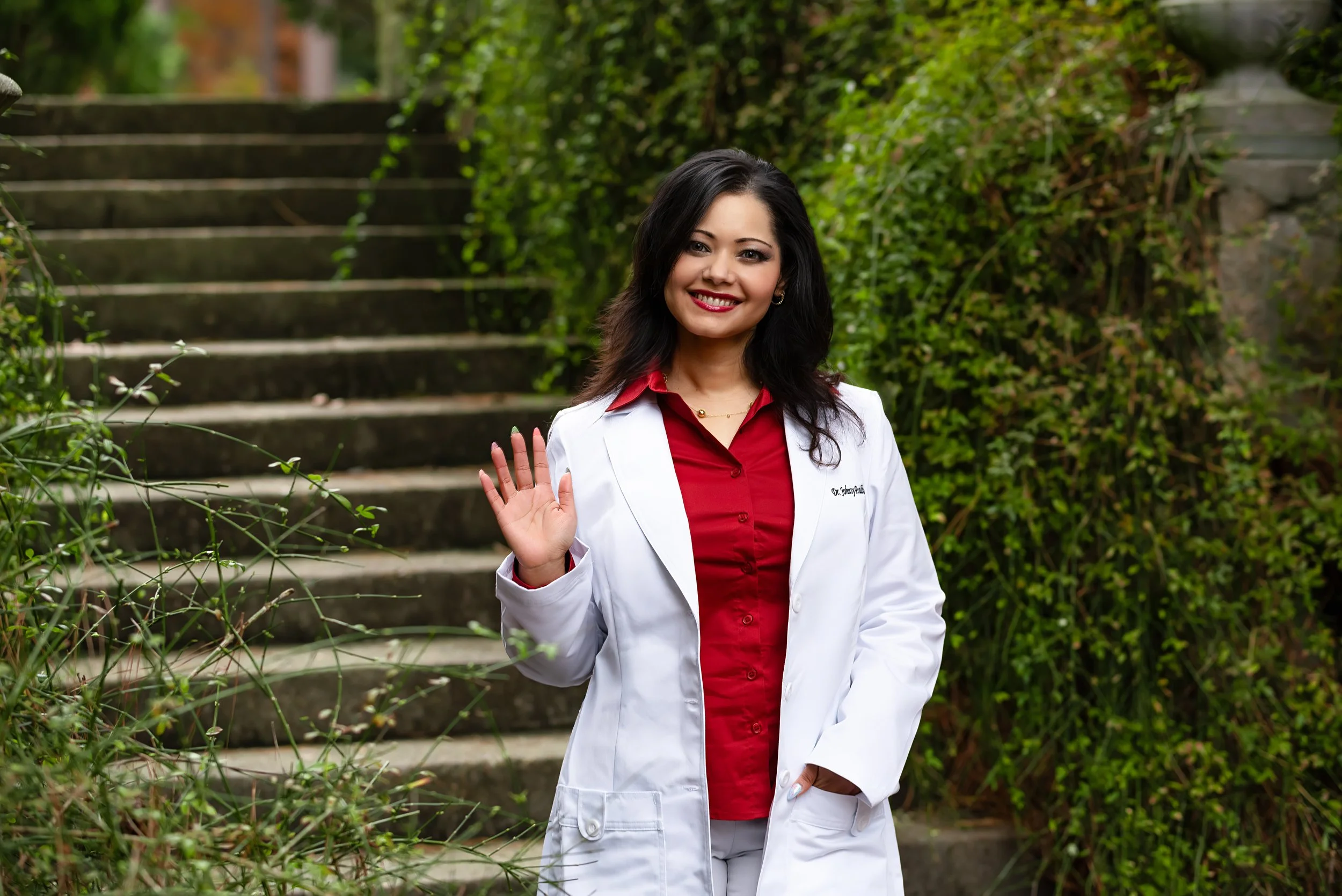 A woman in a white coat waving outdoors near stairs and greenery.