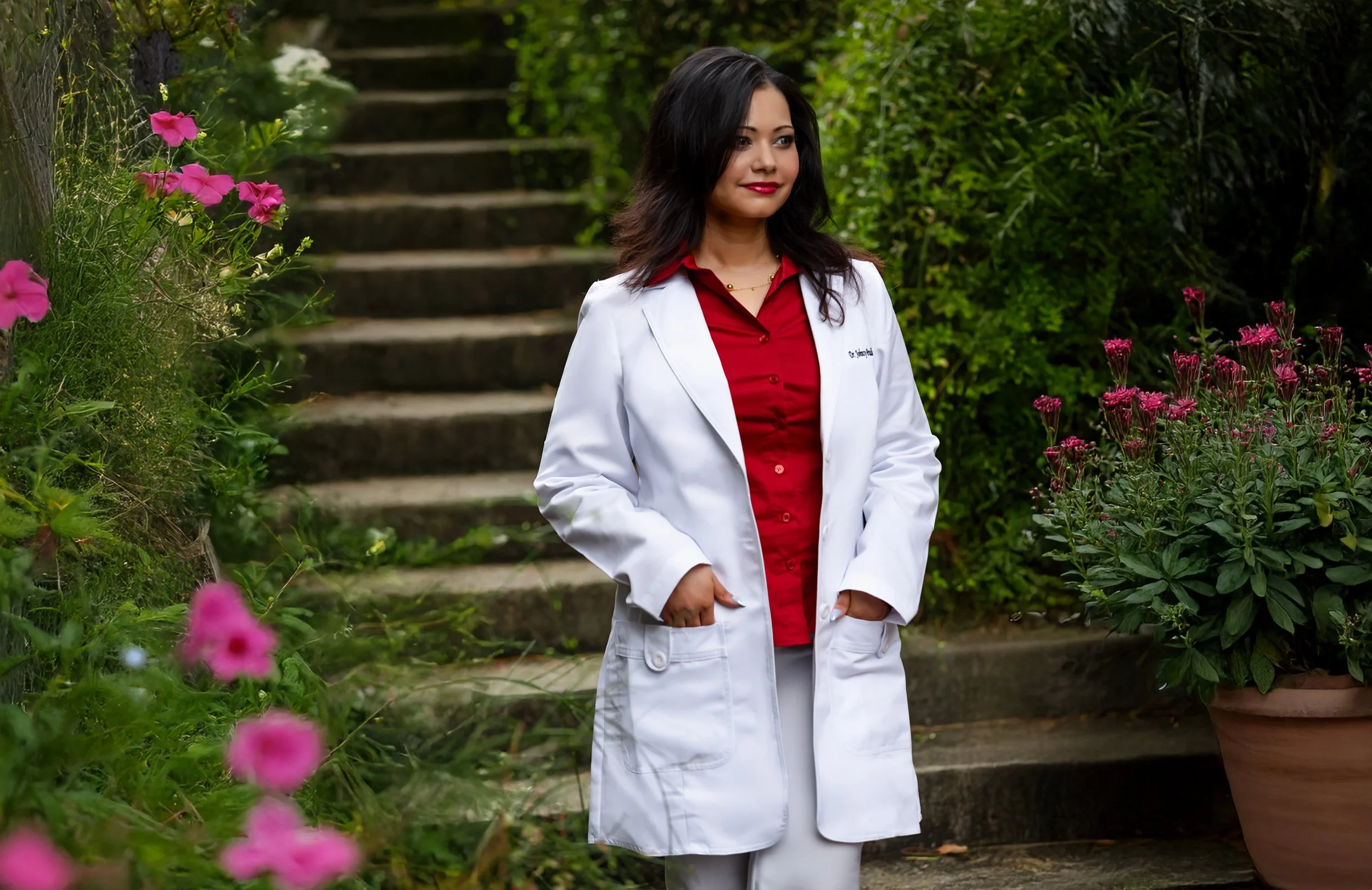 A woman in a white lab coat and red shirt standing outdoors on stone steps surrounded by green and pink flowering plants.