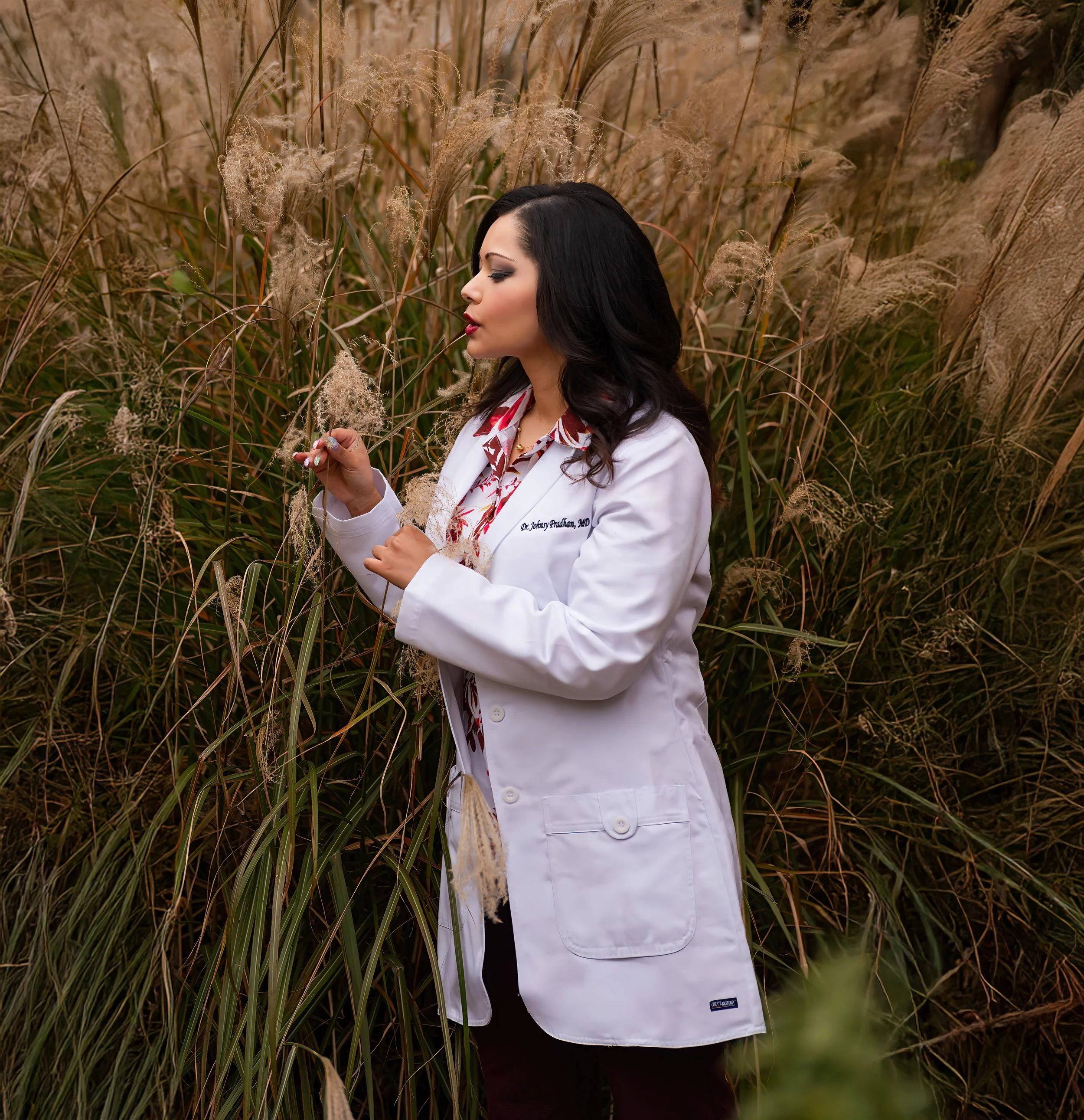 A woman in a white lab coat standing in a field of tall, brown grasses, holding a few stalks of grass and looking at them thoughtfully.