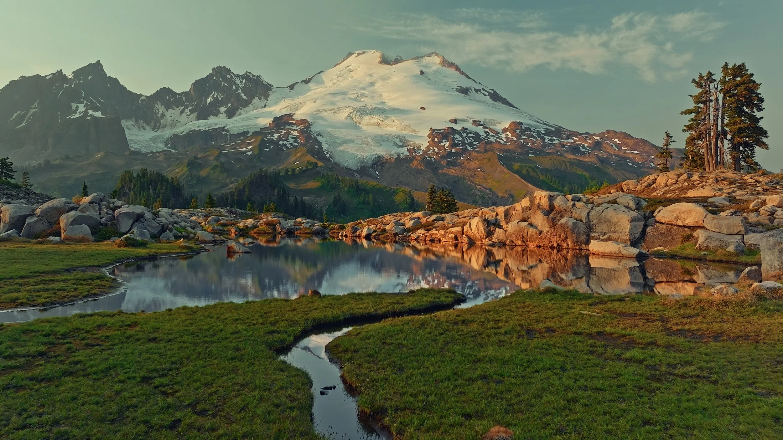 Snow-capped mountain reflecting in a small lake, surrounded by rocks, green grass, and trees.